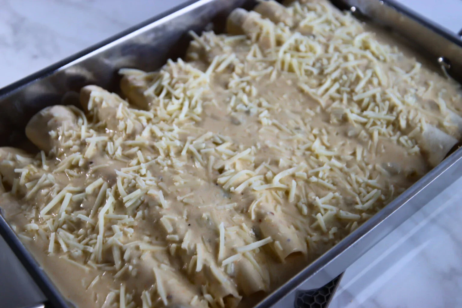 2025-04-26-18.01.40-1536x1024.webp Rolled chicken enchiladas covered with creamy white sauce and shredded Monterey Jack cheese in a baking pan.
